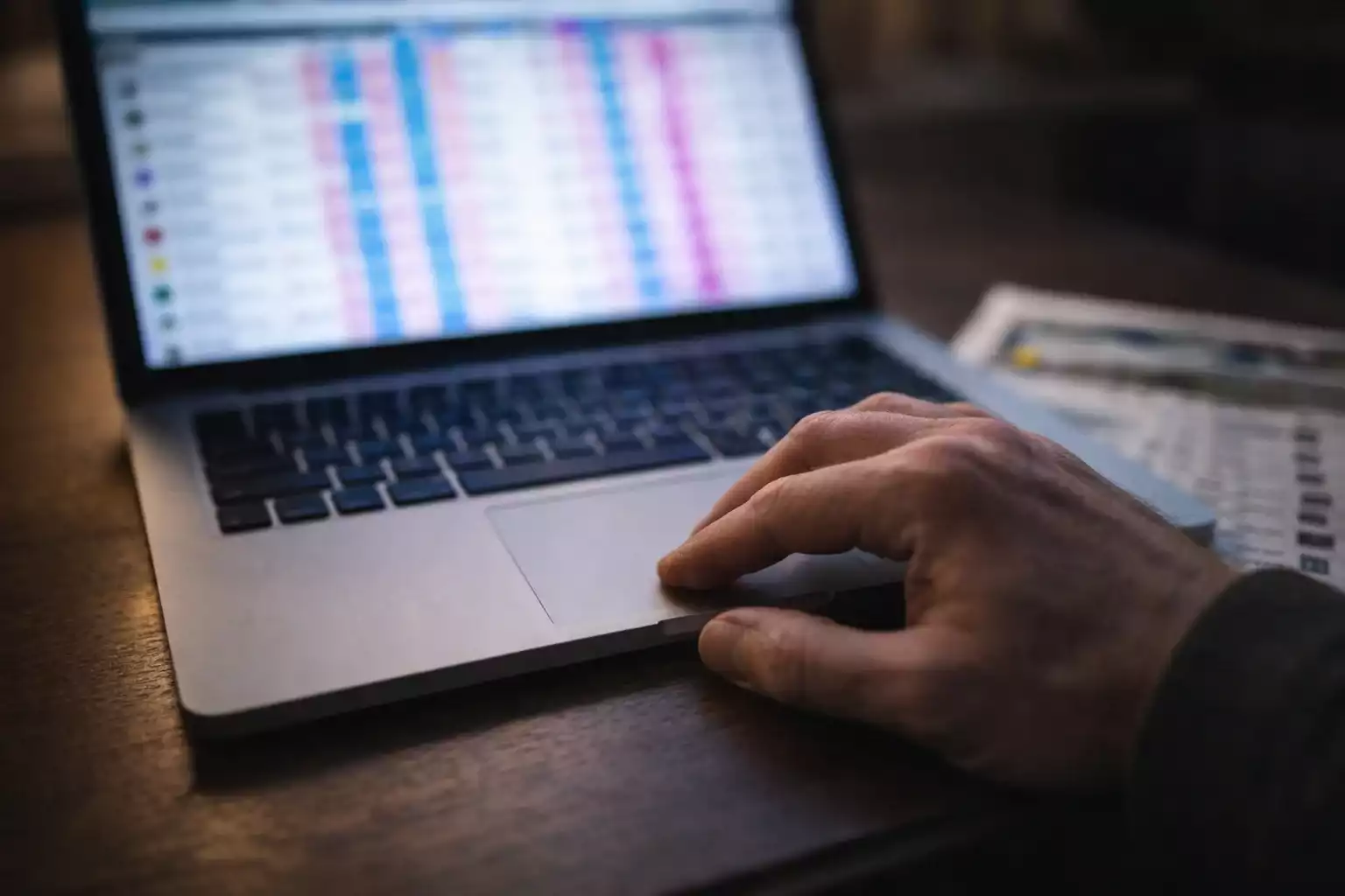 A person's hand using a laptop trackpad with blurred pink and blue exchange columns on screen, a greyhound racecard beside the computer.